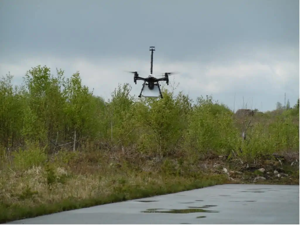 The image shows a quadcopter hovering with the white Aeris sensor attached underneath. The quadcopter also carries a Sparvio sensor system that synchronizes data from Aeris with readings from the TriSonica Wind & Weather Sensor, GPS and other sensors. Sparvio logs all data and transmits it to a ground station to visualize on a map in real-time. In the end, the combination of all data will be used to calculate not only the location of methane emission sources, but also the quantity of gas flow.